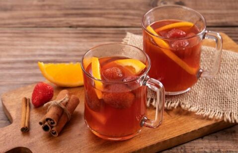 duas canecas de quentão decorados com morango e laranja, paus de canela, servido sob tábua de madeira