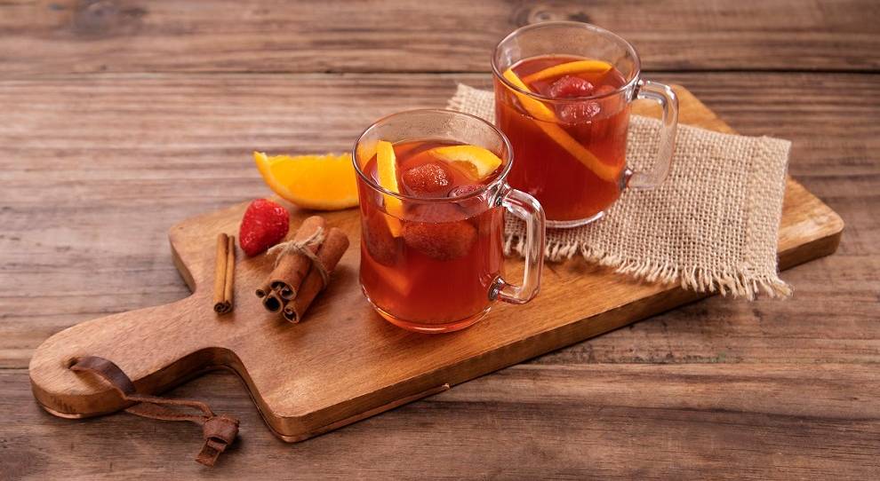 duas canecas de quentão decorados com morango e laranja, paus de canela, servido sob tábua de madeira