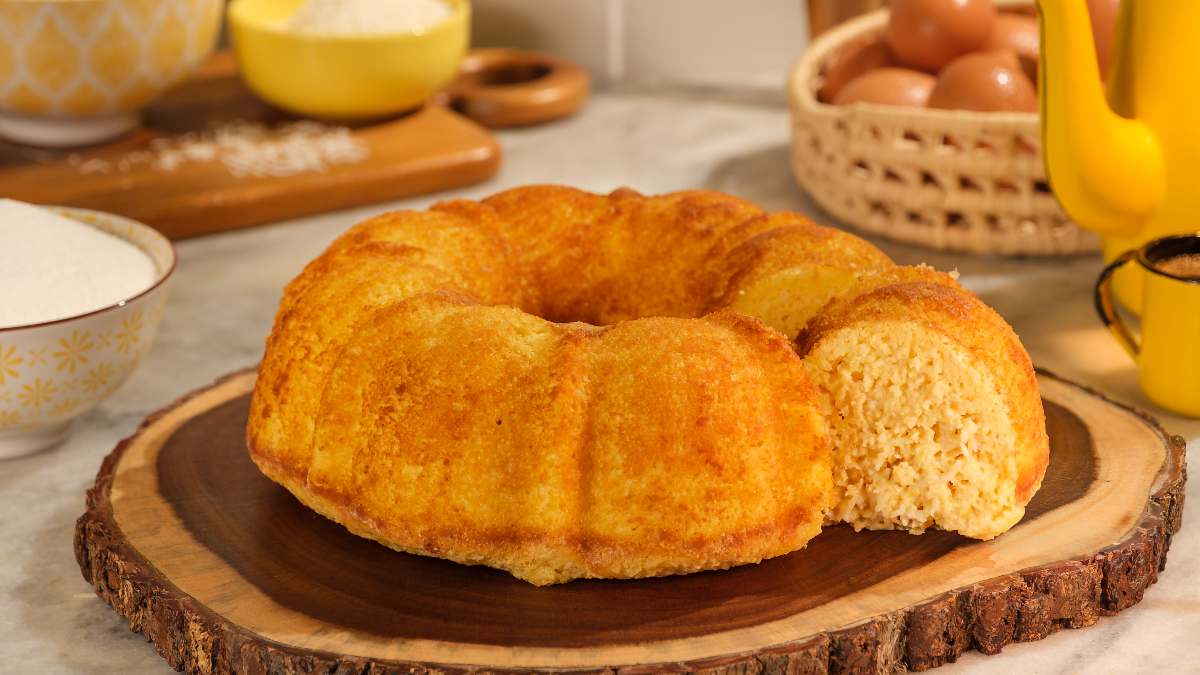 Bolo de milho servido em uma tábua de madeira rústica parecida com um tronco de árvore. O bolo foi cortado e há uma fatia na direita mostrando o interior do bolo, que é meio amarelado e visualmente fofinho. Ao fundo da imagem, do lado esquerdo, uma garrafa de café antiga esmaltada de cor amarela e uma xícara esmaltada também amarela. Mais atrás, uma cesta com ovos caipiras. Do lado esquerdo, uma tábua de madeira com uma tigela amarela redonda com coco ralado e outra maior com farinha de trigo. Mais à frente, do lado esquerdo do bolo, uma tigela com açúcar refinado.