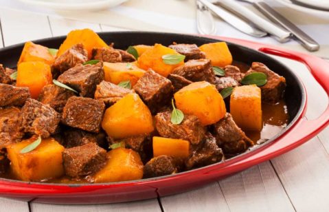 Carne de panela servida com batatas em uma travessa vermelha rasa, em uma mesa de tábuas de madeira branca.