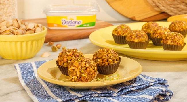 A imagem destaca três unidades de brigadeiro de pistache dispostos em um prato amarelo redondo pequeno que está por cima de um pano de prato azul com listras brancas espaçadas e dobrado. Ao fundo da imagem, do lado direito, outro prato amarelo maior com 6 unidades de brigadeiro de pistache. Do lado esquerdo, em uma tigela amarela, grãos de pistache com casca e no meio da imagem, uma embalagem de Doriana com Sal.