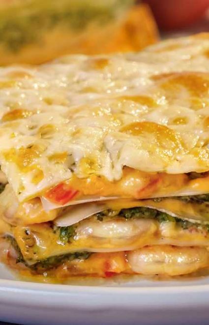 Fatia de lasanha recheada com camarão e molho pesto de coentro servida em um prato redondo branco e azul. Atrás do prato, a travessa com o restante da lasanha de camarão.