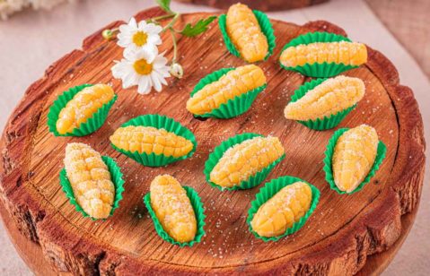A imagem detaca uma porção de brigadeiro de milho em formato de espiga em forminhas verdes imitando a palha, dispostas em uma tábia de madeira que imita um tronco de árvora. Entre os brigadeiros, duas margaridas brancas para decorar.