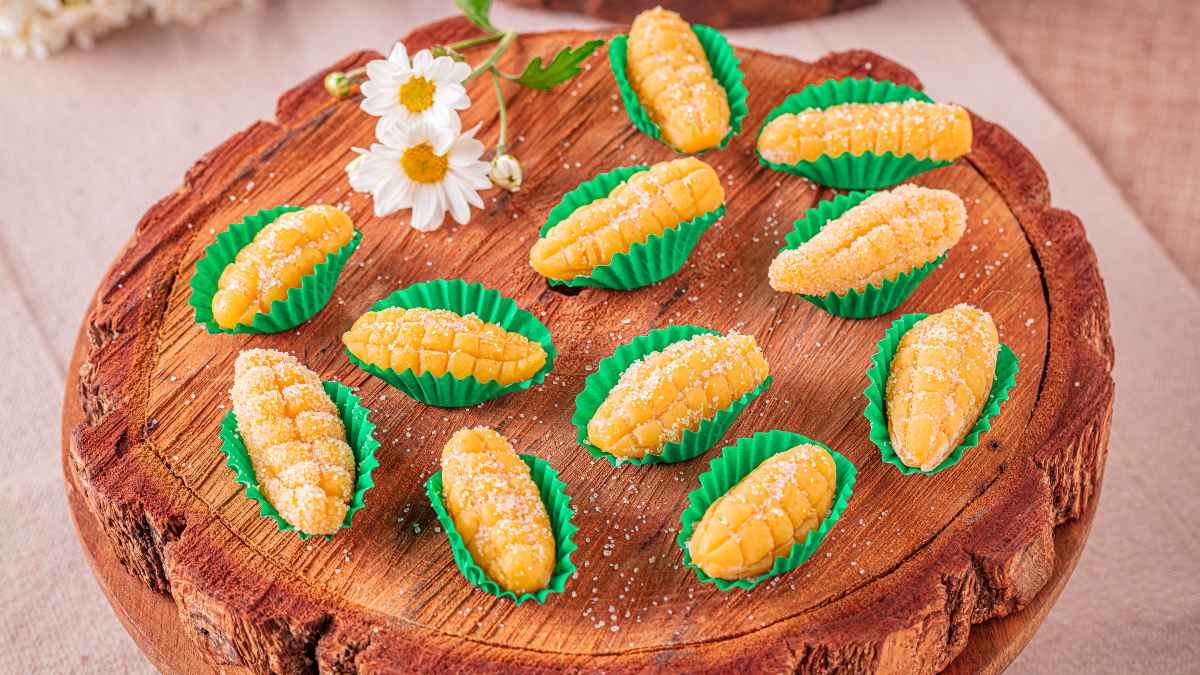 A imagem detaca uma porção de brigadeiro de milho em formato de espiga em forminhas verdes imitando a palha, dispostas em uma tábia de madeira que imita um tronco de árvora. Entre os brigadeiros, duas margaridas brancas para decorar.