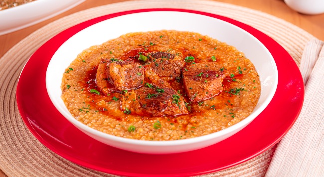 A imagem destaca um prato branco fundo com uma porção de canjiquinha com costelinha. O prato estpa sobre um outro prato maiol, raso e vermleho