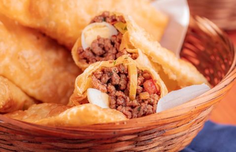 Hambúrguer gourmet de carne bovina com cebola crispy, bacon crocante, maionese e folhas de rúcula servido no pão brioche. Ao lado, um molho agridoce tipo barbecue. Ao fundo, uma taça de vinho tinto.