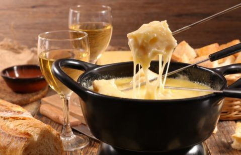 Imagem destaca o fondue de queijo posto sobre a mesa com duas taças de vinho branco