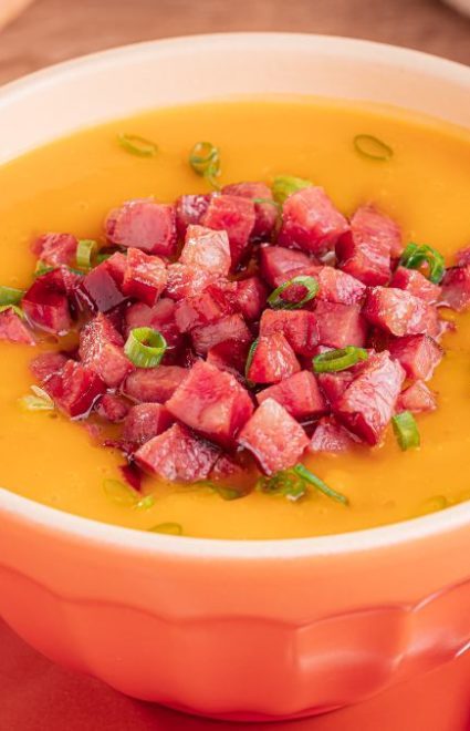 A imagem destaca uma tigela branca de caldo de abóbora com linguiça calabresa picada. A tigela está sobre um aparador de prato laranja. Do lado direito, uma colher prateada.