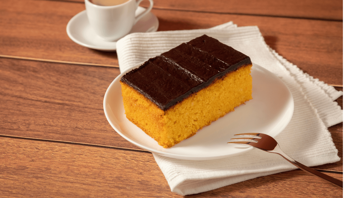 A imagem mostra uma fatia de bolo de cenoura com cobertura de chocolate durinha, servida em um prato branco sobre um guardanapo de tecido na mesma cor. A fatia de bolo tem uma coloração laranja vibrante, típica do bolo de cenoura, com uma cobertura espessa e brilhante de chocolate que cobre toda a superfície superior do bolo. Ao lado do prato, há um garfo prateado. No fundo, sobre uma superfície de madeira, pode-se ver uma xícara de café preto em uma pequena xícara branca, complementando a cena de um lanche ou café da tarde. O contraste das cores do bolo, prato e guardanapo com a superfície de madeira e a xícara de café traz uma sensação acolhedora e reconfortante.