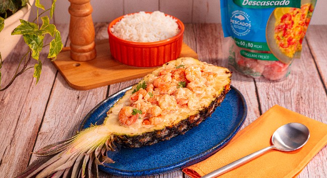 Camarão ao molho de coco servido no abacaxi inteiro, cortado ao meio, e disposto em um prato azul escuro. Ao fundo, uma tigela com arroz branco.