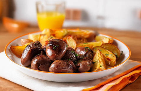 Um prato com corações de galinha e batatas assadas, em destaque sobre uma mesa rústica de madeira. A refeição, com cores vibrantes e aspecto apetitoso, convida para uma degustação caseira. Um copo com suco de laranja e outros detalhes culinários completam a cena.