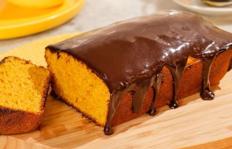 Um bolo de cenoura com cobertura de chocolate está em foco, com uma fatia separada do resto do bolo. O bolo em si tem uma cor laranja vibrante, com a textura de um bolo de cenoura clássico. A cobertura de chocolate é rica e brilhante, com uma pequena porção derramada em um padrão irregular.