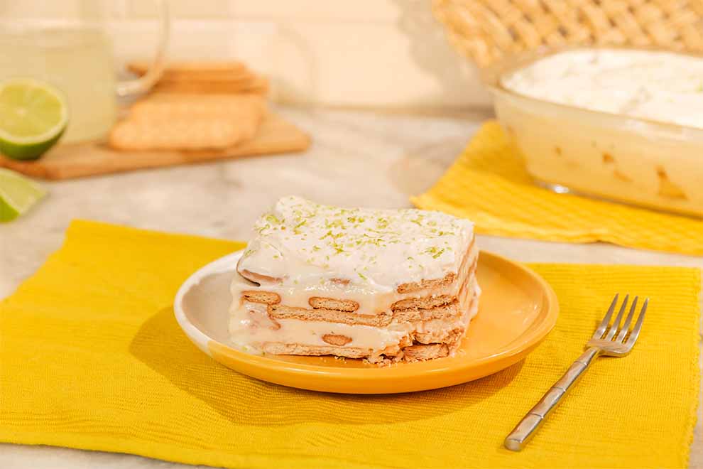 um pedaço de pavê de limão dentro de um prato redondo de porcelana em branco e amarelo com um garfo ao lado em cima de um pano amarelo