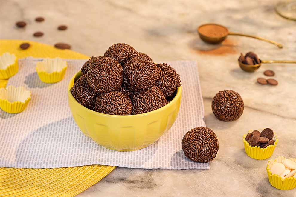 um pequeno bowl amarelo com brigadeiros dentro e com formas pequenas de papel para brigadeiro em volta em cima de um guardanapo listrado acinzentado e de um jogo americano de palha