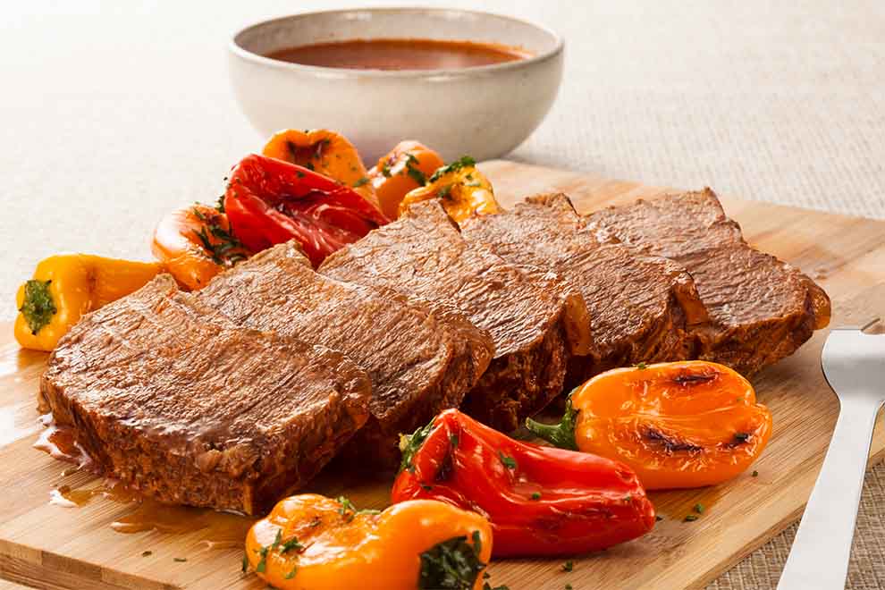 contra-filé assado com legumes grelhados em cima de uma tábua de madeira com um garfo de churrasco ao lado e um bowl de molho ao fundo