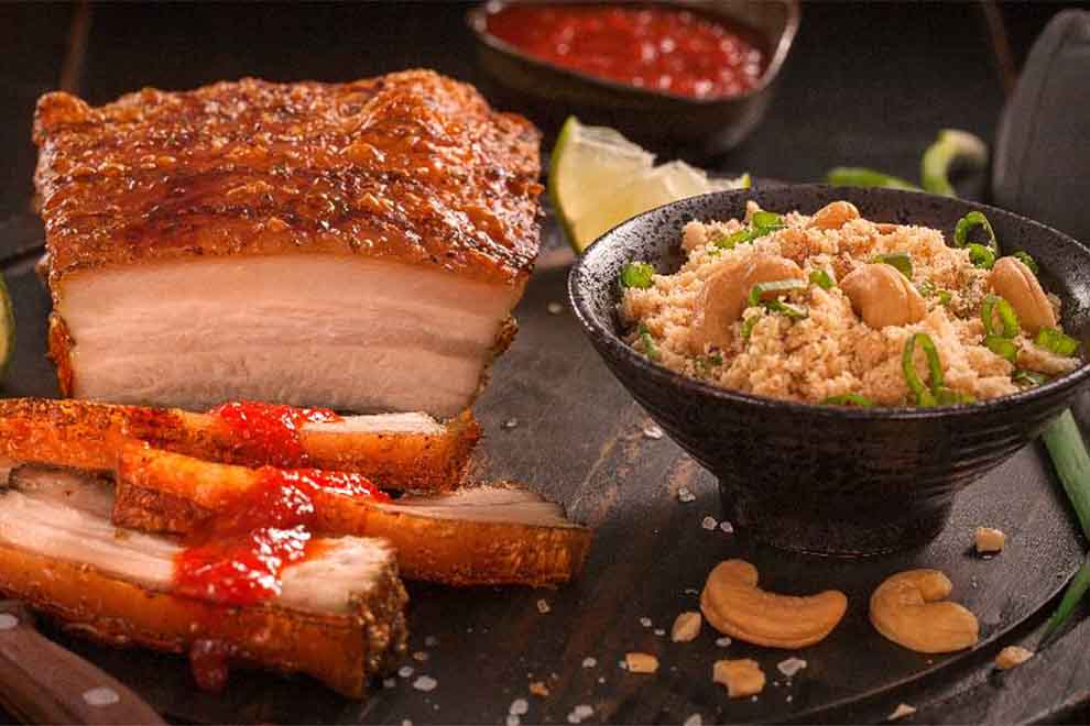 panceta suína com farofa de castanha de caju dentro de um pequeno bowl preto ao molho agridoce dentro de uma base de porcelana preta em cima de uma mesa escura