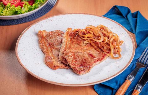 bife acebolado dentro de um prato de porcelana embaixo de um pano azul em cima de uma mesa de madeira