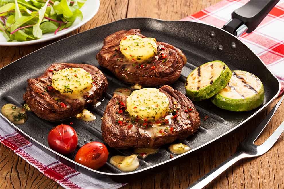 pedaços de contra-filé grelhado com manteiga de ervas por cima e legumes grelhados ao lado, em cima de uma frigideira para grelhar com um garfo de churrasco ao lado e em cima de um guardanapo xadrez vermelho
