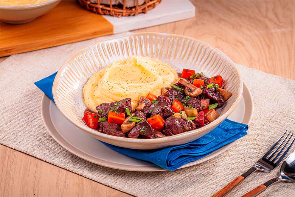 carne de panela ao molho vinho com polenta ao lado dentro de um prato fundo de porcelana bege, em cima de uma base de porcelana bege e com guardanapo azul e talheres ao lado