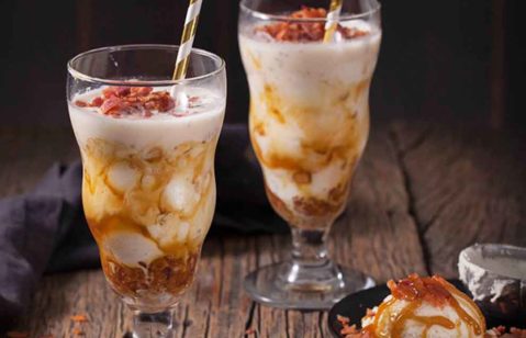 milkshake de creme com caramelo e bacon com canudos listrados em branco e dourado em cima de uma mesa de madeira