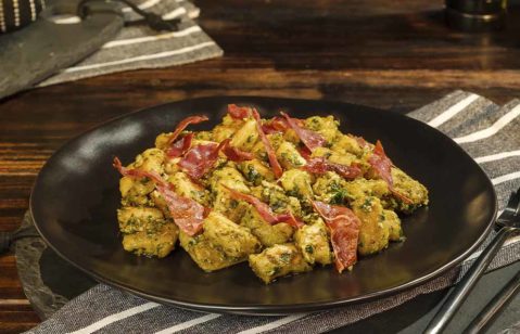 Nhoque de batata doce com molho pesto e presunto crú, em cima de um prato escuro, sob uma mesa de madeira.