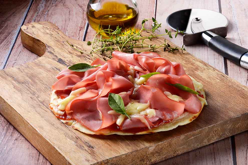 pizza de mortadela de frigideira em cima de uma tábua de madeira com azeite e um pequeno ramo de alecrim ao lado, em cima de uma mesa de madeira