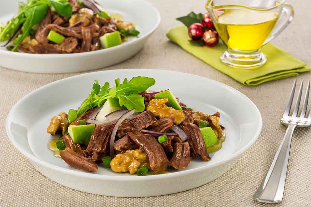 salada de músculo, maçã e nozes dentro de um prato branco de porcelana com um garfo ao lado em cima de uma mesa com jogo americano cinza