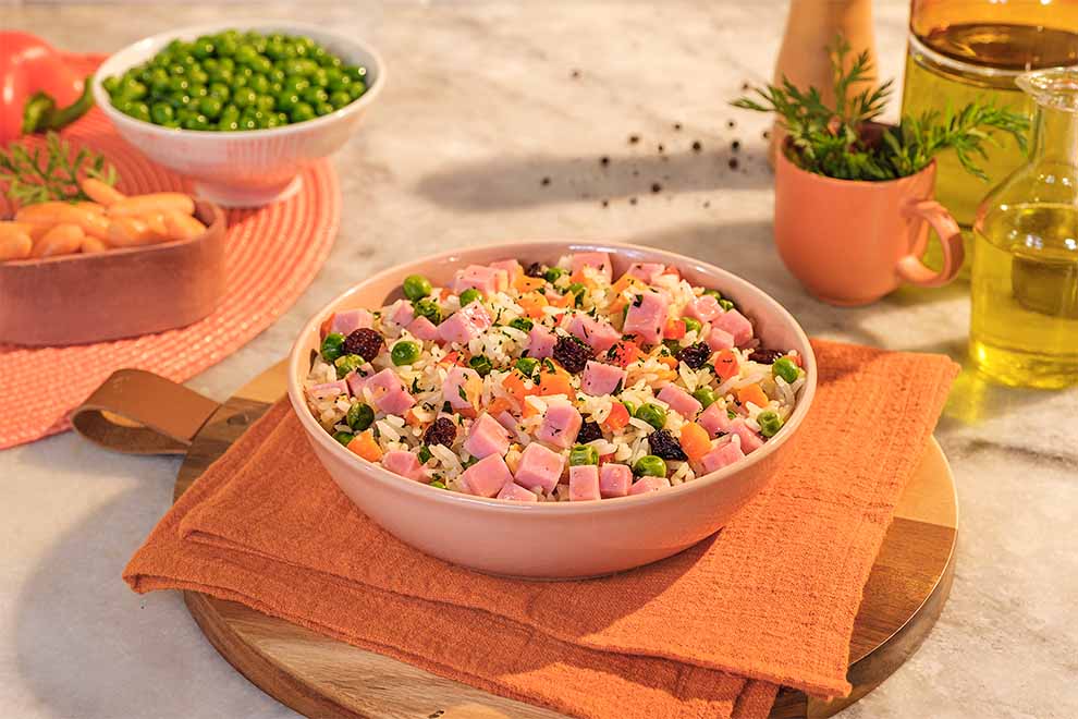 arroz à grega com presunto dentro de um bowl de porcelana rosada em cima de um guardanapo laranja e uma tábua de madeira ao fundo alguns dos ingredientes usados para o preparo e tudo em cima de uma mesa de mármore