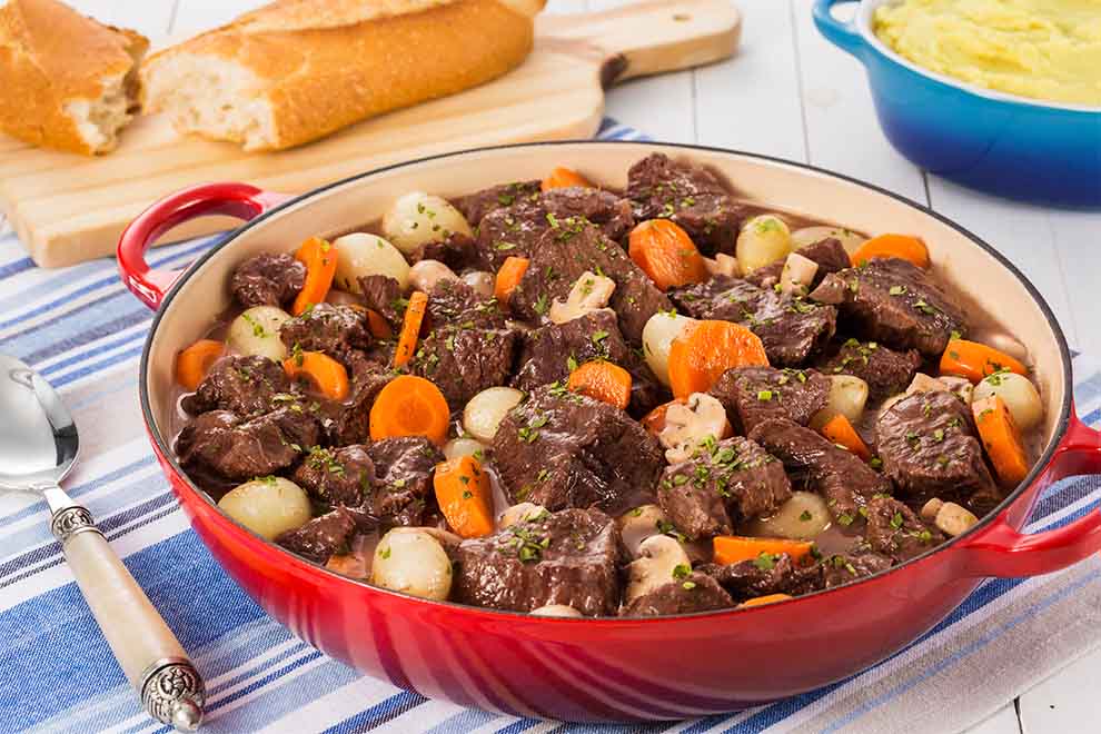 boeuf bourguignon de paleta dentro de uma grande panela avermelhada em cima de um pano listrado em azul e branco em cima da mesa