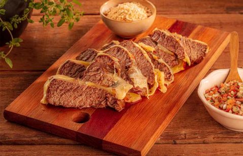 cupim com queijo bem passada em cima de uma tábua de madeira e um pequeno bowl com farofa e em cima de uma mesa de madeira