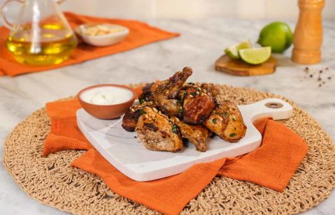 várias coxas de frango feito na airfryer, com um pequeno bowl de molho ao lado e em cima de uma pequena tábua branca de marmore, em cima de um pano alaranjado e de um jogo americano redondo de palha