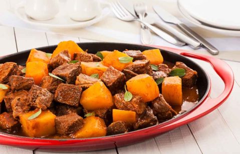 picadinho de carne com mandioca dentro de uma travessa de porcelana preto e vermelho em cima de uma mesa de madeira com talheres ao lado